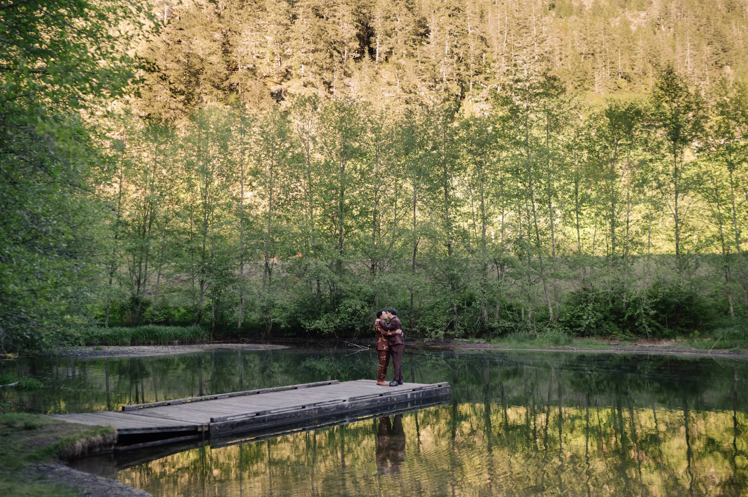 Couple hugging and kissing on a wooden dock by a lake, surrounded by greenery and trees.