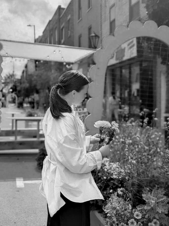 A woman holding a small bouquet of flowers outside a flower shop on a city street.