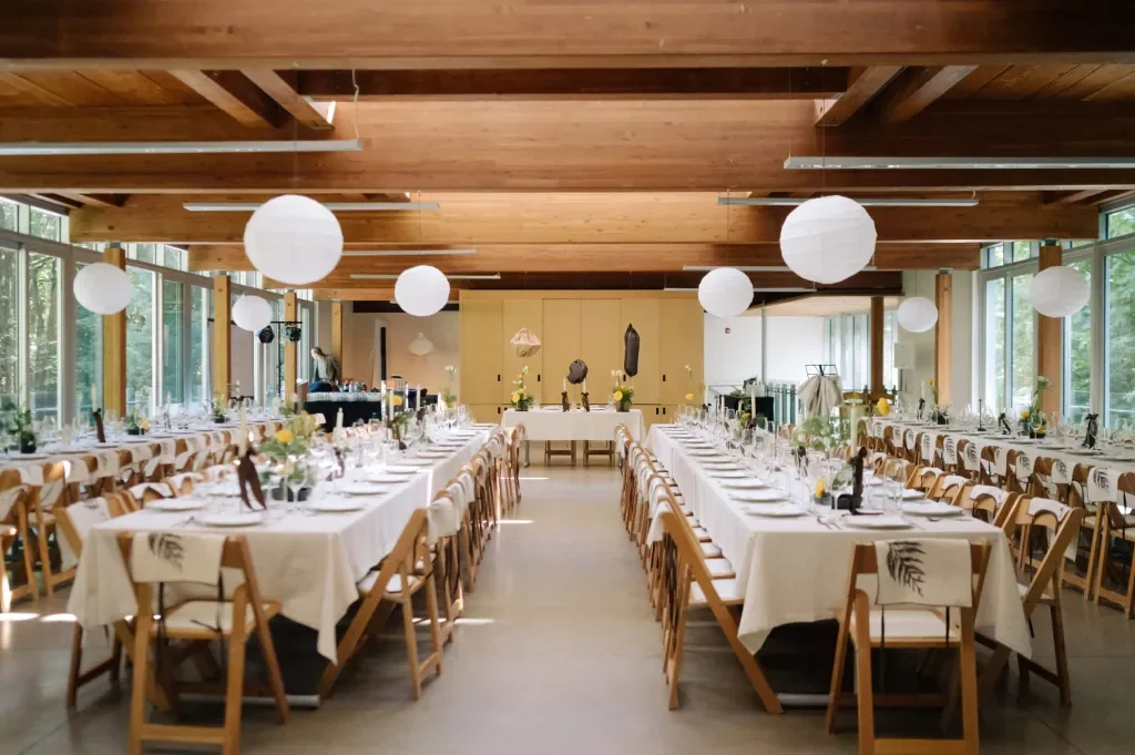 Long dining tables set for a formal event in a bright, wood-paneled room with large windows, white tablecloths, floral centerpieces, and paper lanterns hanging from the ceiling.