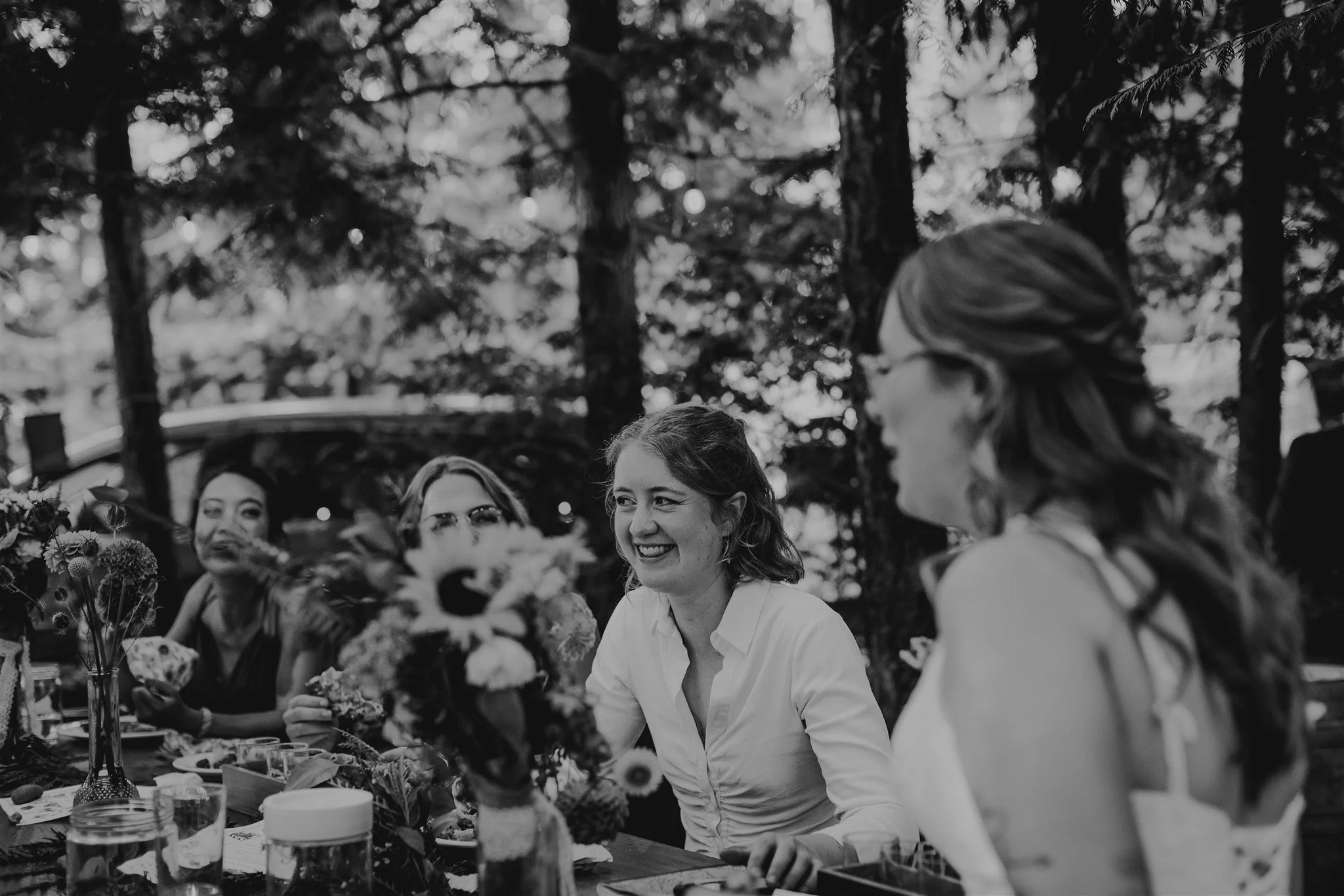 Black and white photo of women sitting at a table outdoors, engaging in conversation and smiling, in a wooded area.