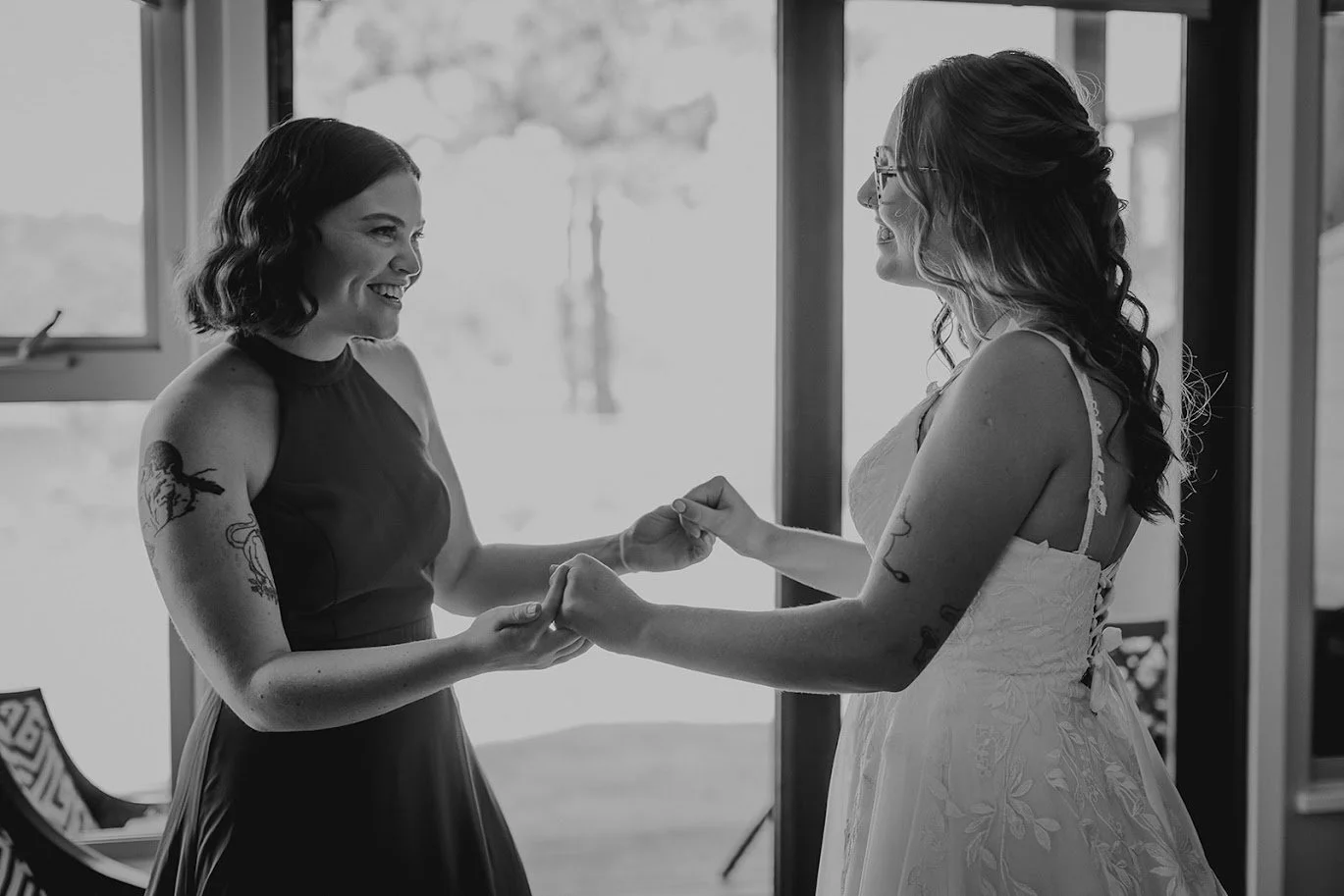 Two women smiling and holding hands indoors near a window, one in a dark dress and the other in a white dress, engaging in a meaningful moment.