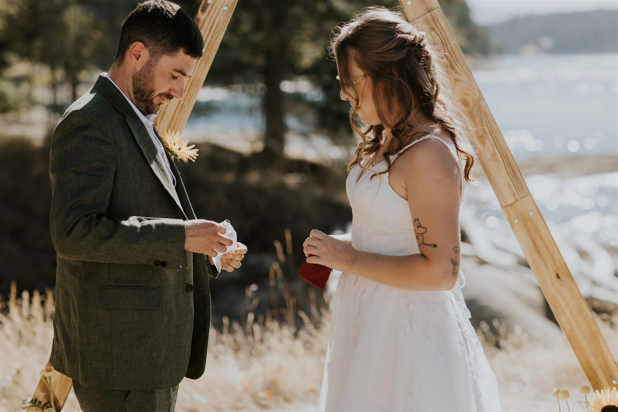 A couple getting married outdoors on a sunny day near the water, with a wooden wedding arch behind them.