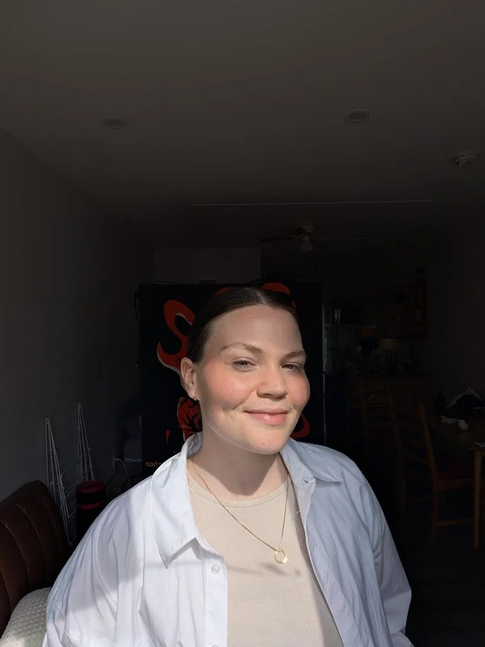 A young woman with dark hair pulled back, smiling, wearing a white shirt over a beige top and a gold necklace, standing in a room with a dark background.
