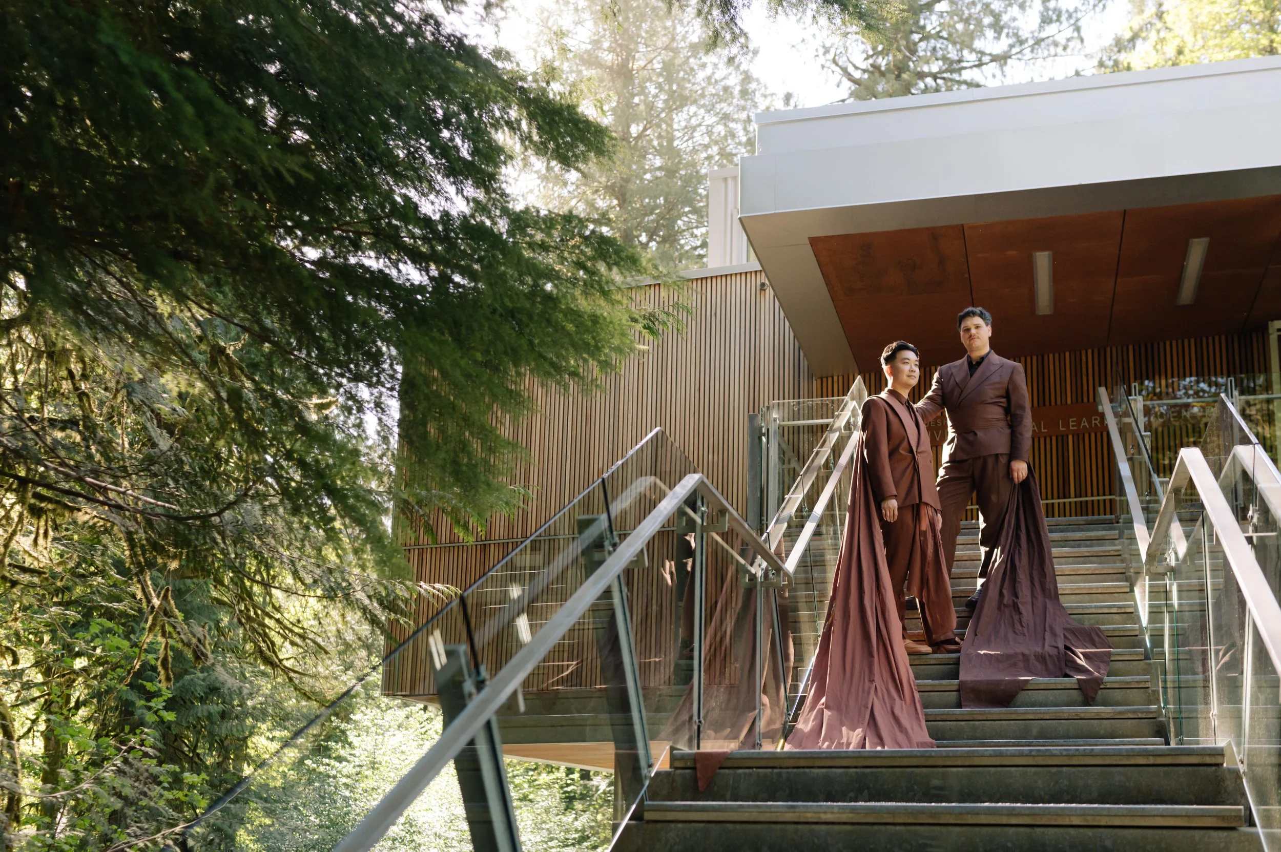 Two models dressed in brown formal attire stand on an outdoor staircase with glass railings, surrounded by trees and modern building architecture.