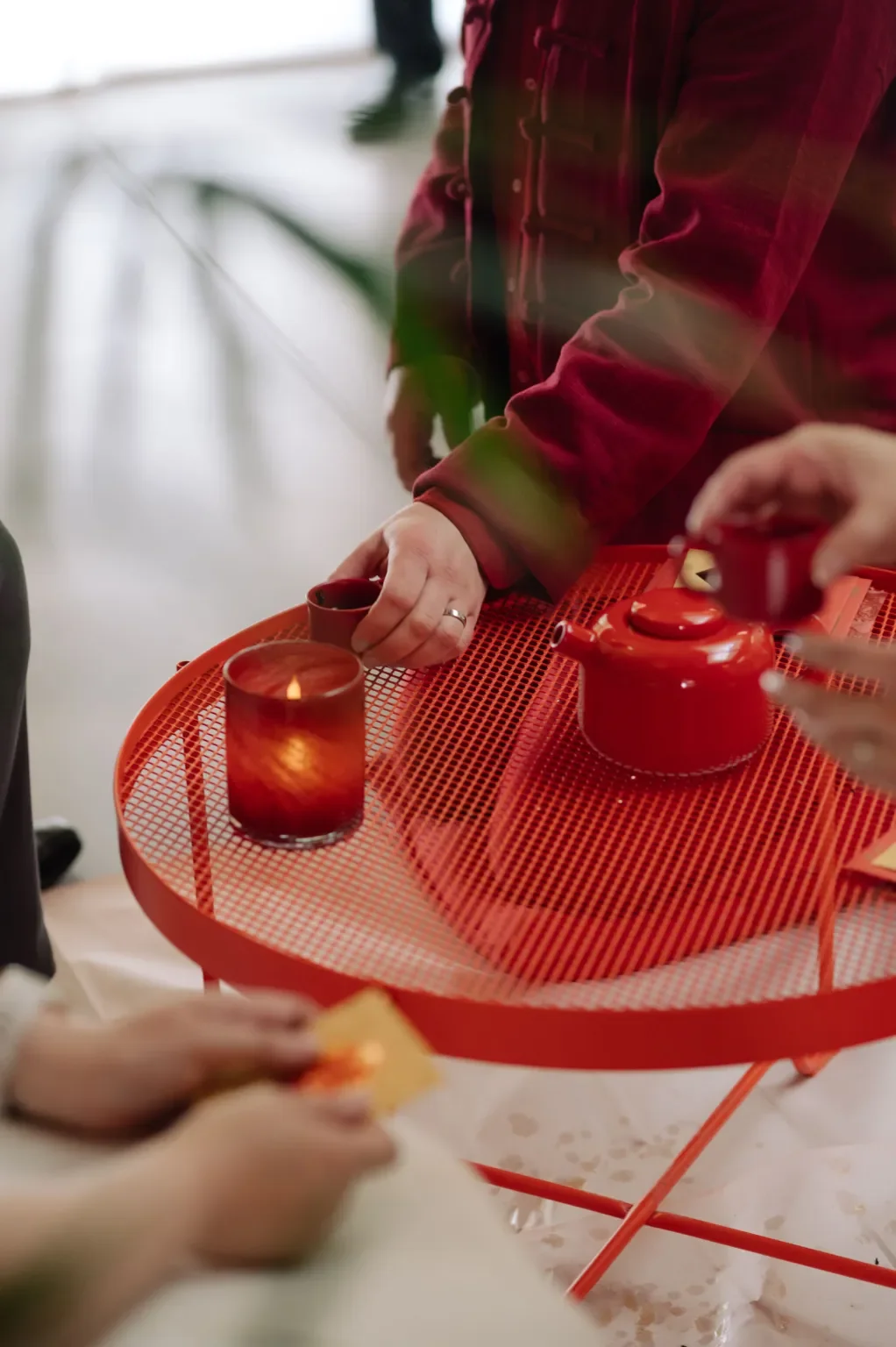 Person in a red jacket pouring tea into cups on a red metal table, with candles and food nearby.