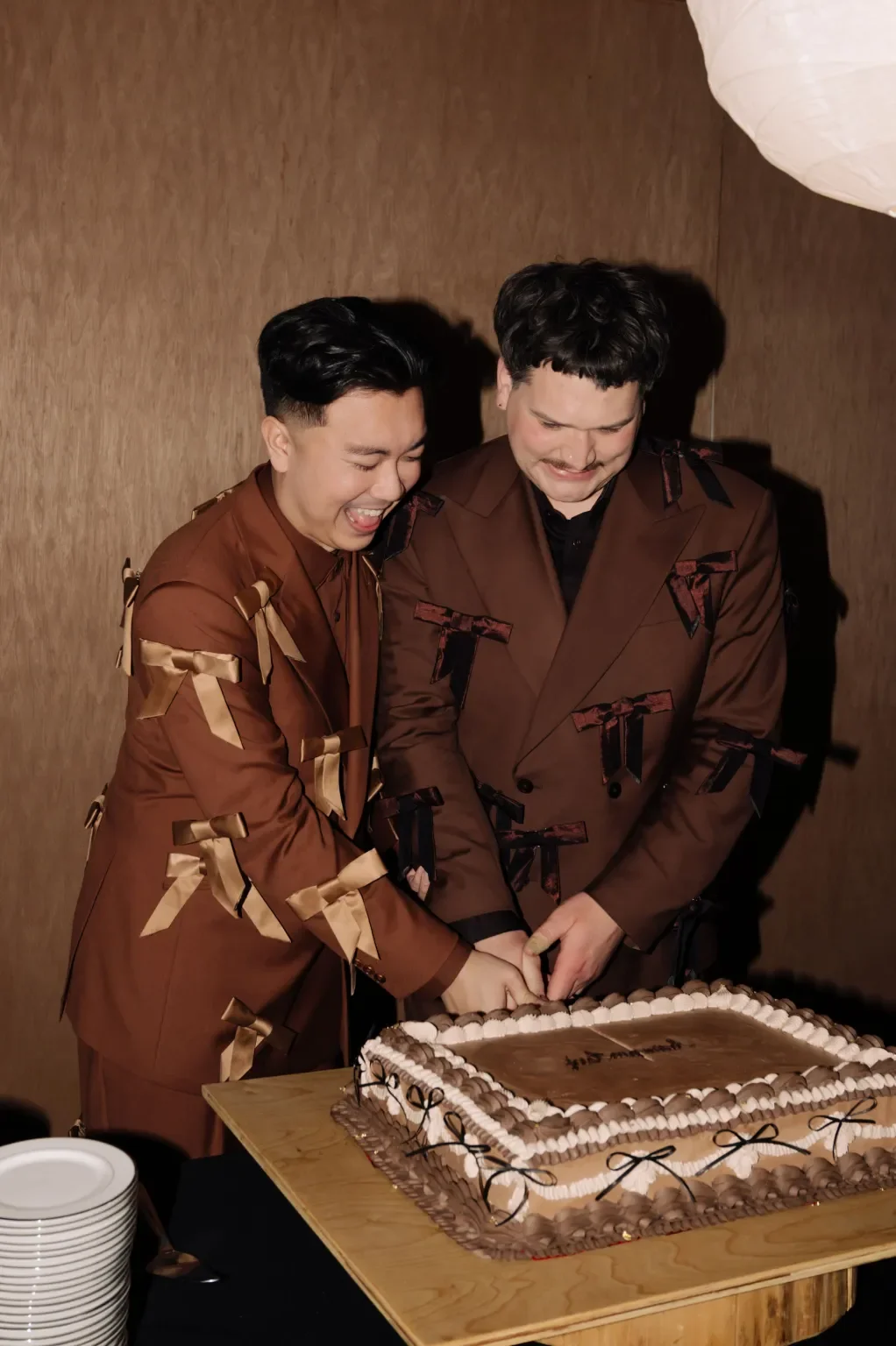Two men dressed in brown suits with ribbon decorations cut a cake together. They are smiling and appear happy, standing in front of a wooden wall.