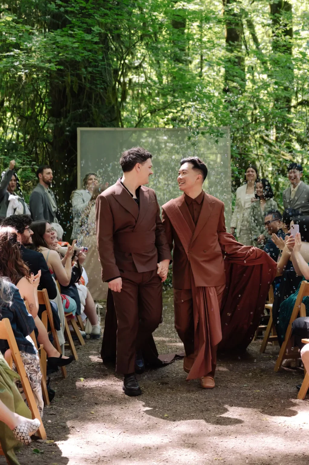 Two men in brown suits holding hands, walking down the aisle during a wedding ceremony in a forest, surrounded by seated guests and smiling onlookers, with trees and sunlight in the background.