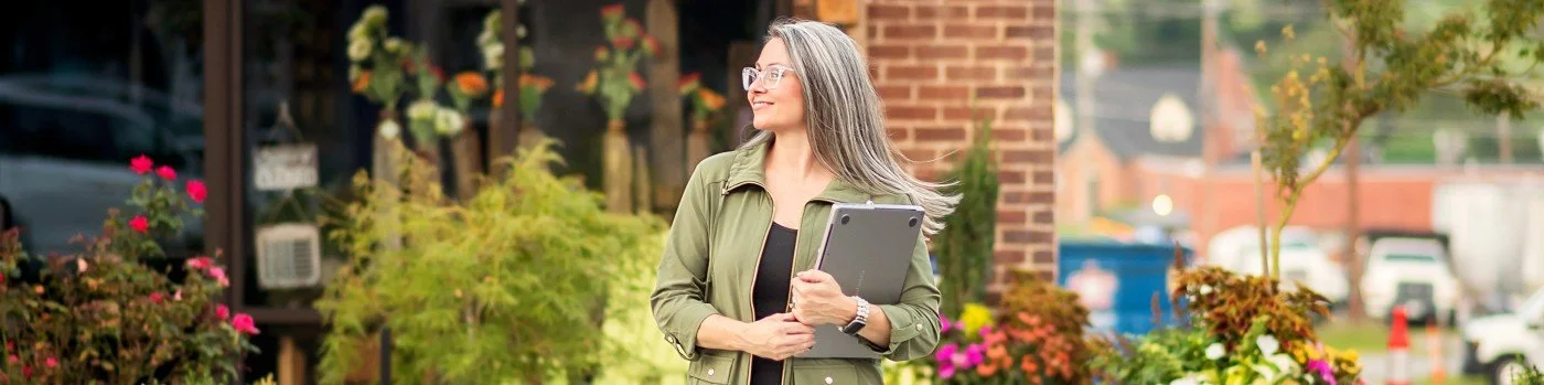 A woman with long gray hair and glasses holding a tablet, standing outdoors in a quaint downtown with trees and buildings in the background.