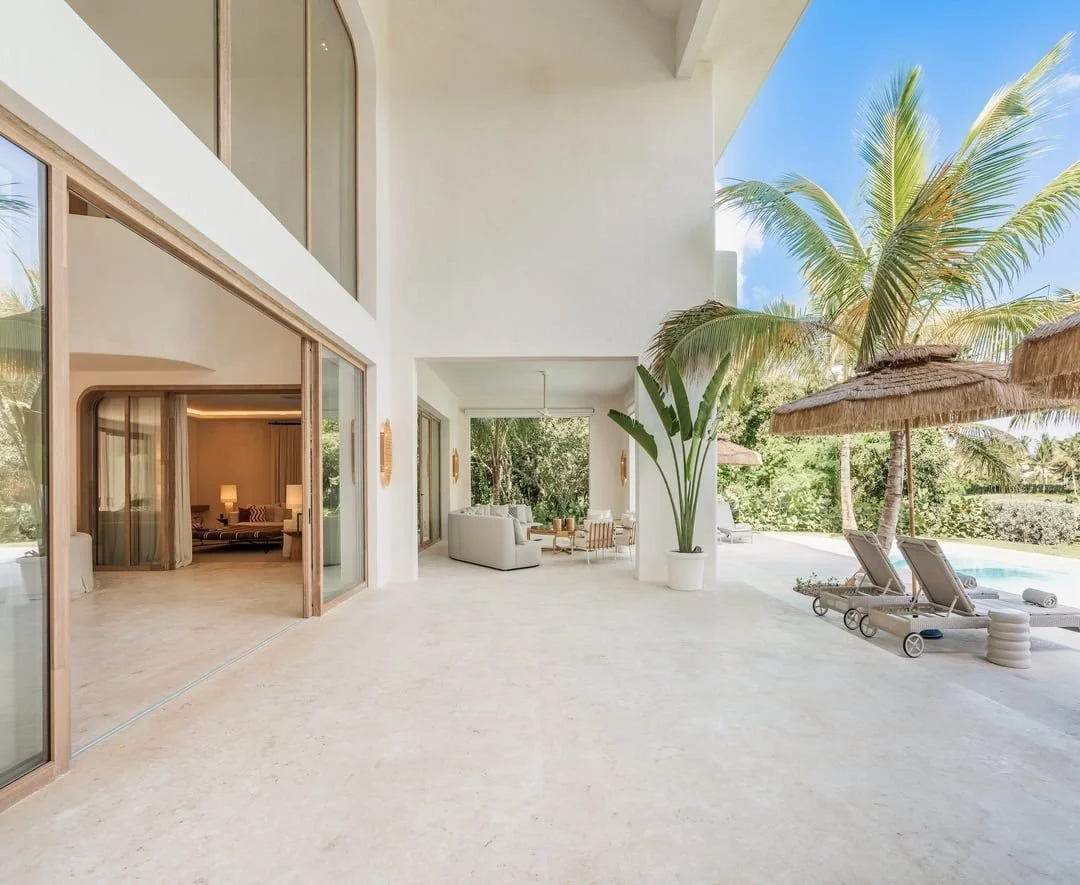 Terraza de casa con muebles blancos, plantas tropicales y vista a jardín y piscina con sombrillas de paja.
