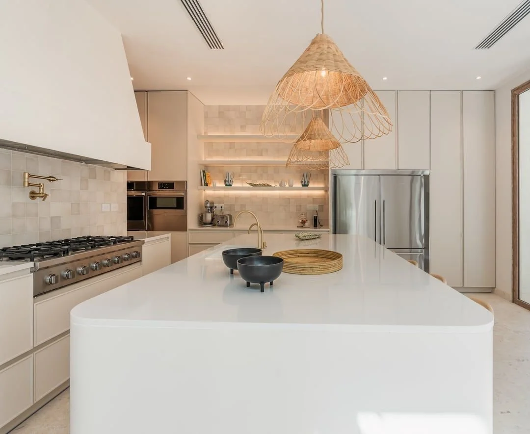 Cocina moderna y luminosa con isla central blanca, lámparas de mimbre colgantes, electrodomésticos de acero inoxidable y estanterías con decoraciones.