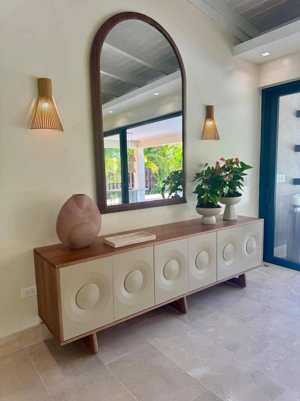 Un mueble de madera con puertas blancas y círculos decorativos, decorado con una vasija de cerámica y dos macetas con plantas, un espejo en la pared y de fondo una vista de un patio exterior.