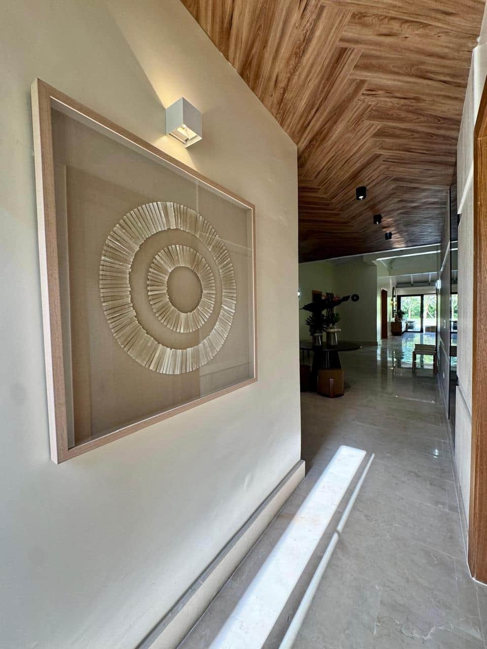 Pasillo interior con pared pintada de color claro, decorada con un marco de madera y un arte en relieve en forma de círculos. El pasillo tiene un techo de madera y paredes, con iluminación empotrada y una iluminación adicional en la parte superior de la pared. La luz natural entra por grandes ventanas al fondo.