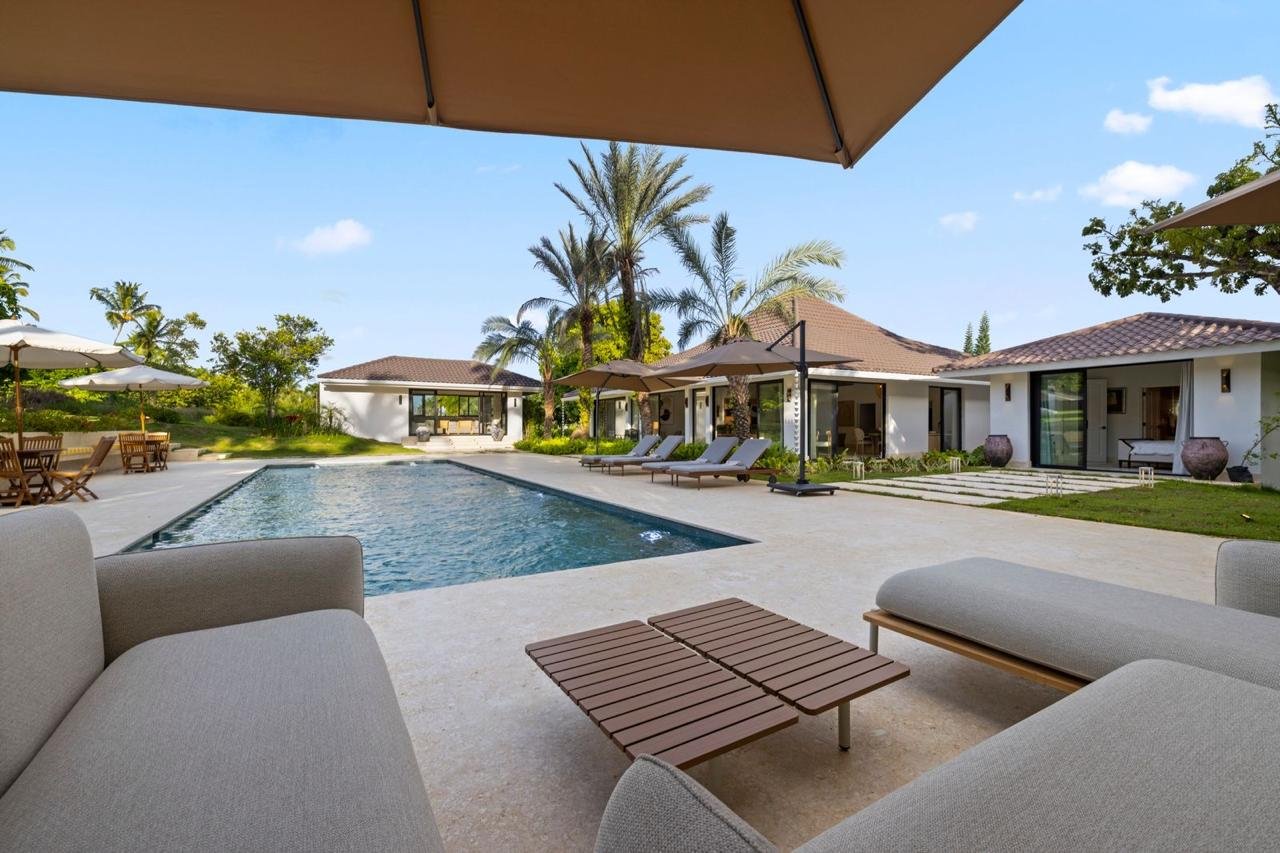 Vista de una piscina en un villa de lujo con mobiliario exterior, sombrillas y casas modernas, rodeada de palmeras y vegetación, en un día soleado con cielo despejado.