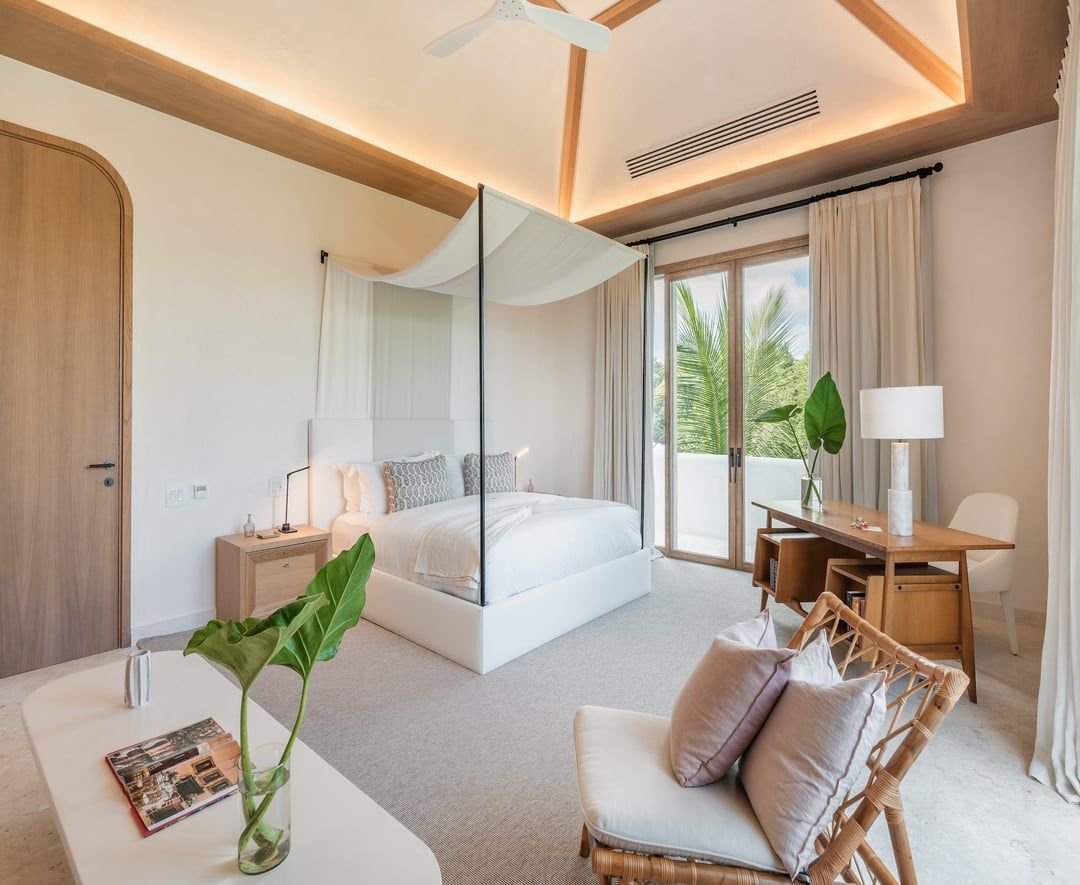 Habitación moderna con cama con dosel de cortinas blancas, escritorio de madera, silla de mimbre con cojines, balcón con vista a plantas verdes y palmeras, y lámparas de mesa y de pie.