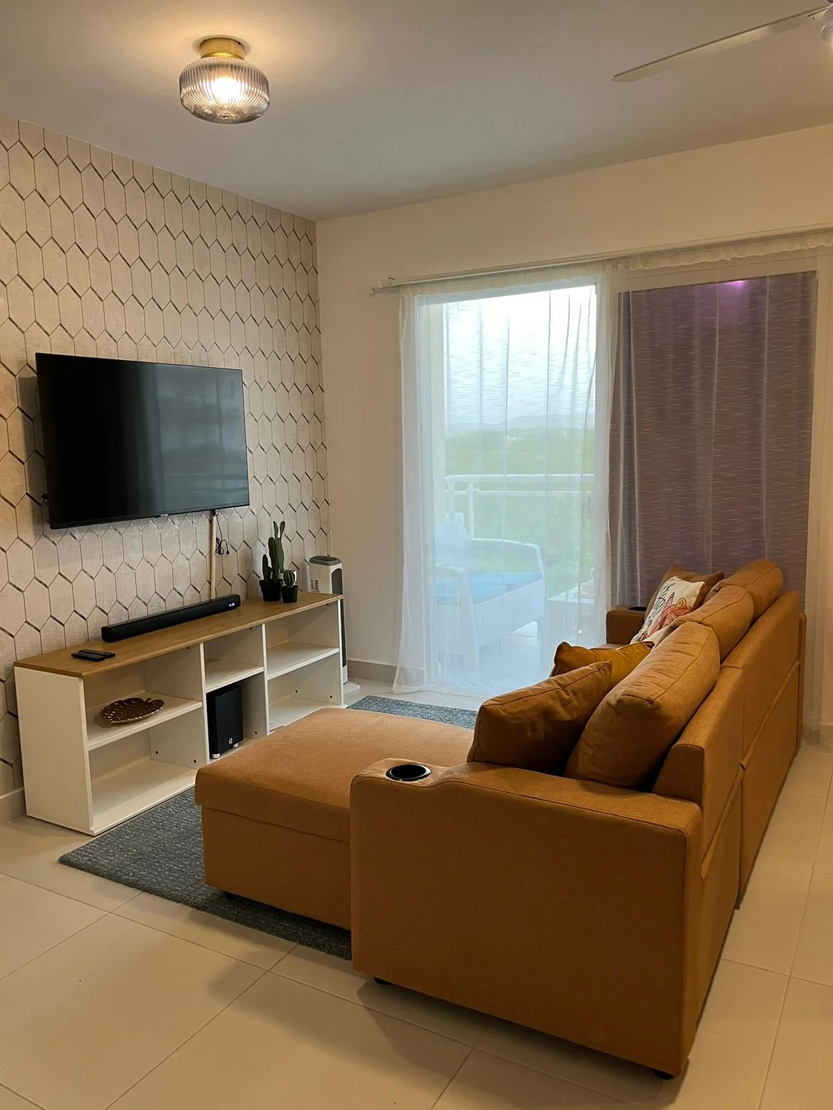 Salón con sofá color mostaza, televisor en la pared, mesa de entretenimiento con decoraciones, cortinas y puerta de balcón con vista a una terraza.