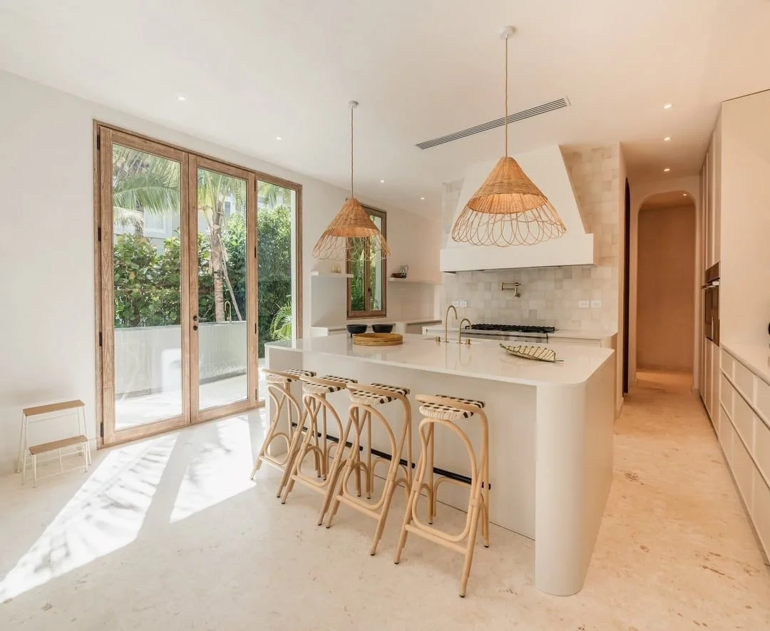 Cocina moderna con isla blanca, asientos de mimbre, lámparas de mimbre, paredes blancas, ventanas grandes con vista a plantas tropicales.