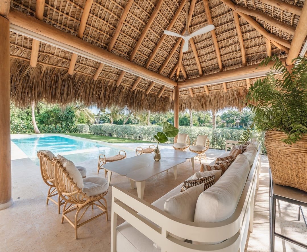Área de descanso exterior con muebles de madera y cojines bajo una palapa de palma, cerca de una piscina y rodeada de vegetación tropical.