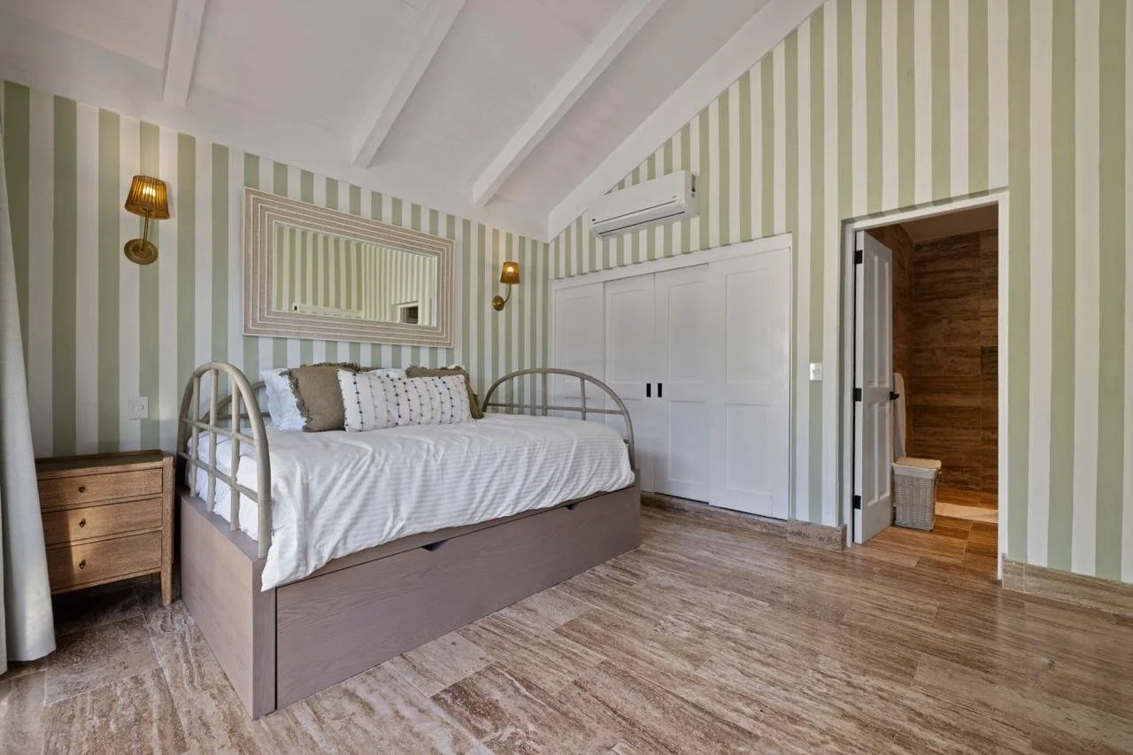 Habitación con paredes de rayas verdes y blancas, cama con cabecera de metal, espejo grande en la pared, lámparas de pared, aire acondicionado, suelo de piedra y puerta a un baño con paredes de madera.