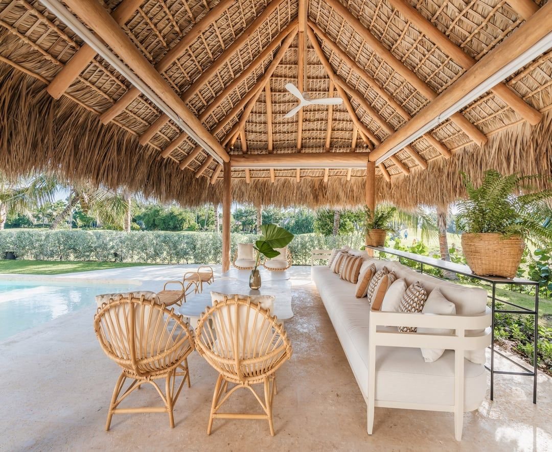 Área de descanso al aire libre con techo de paja y muebles de mimbre, sillones y una larga banca con cojines, rodeada de vegetación y cerca de una piscina.