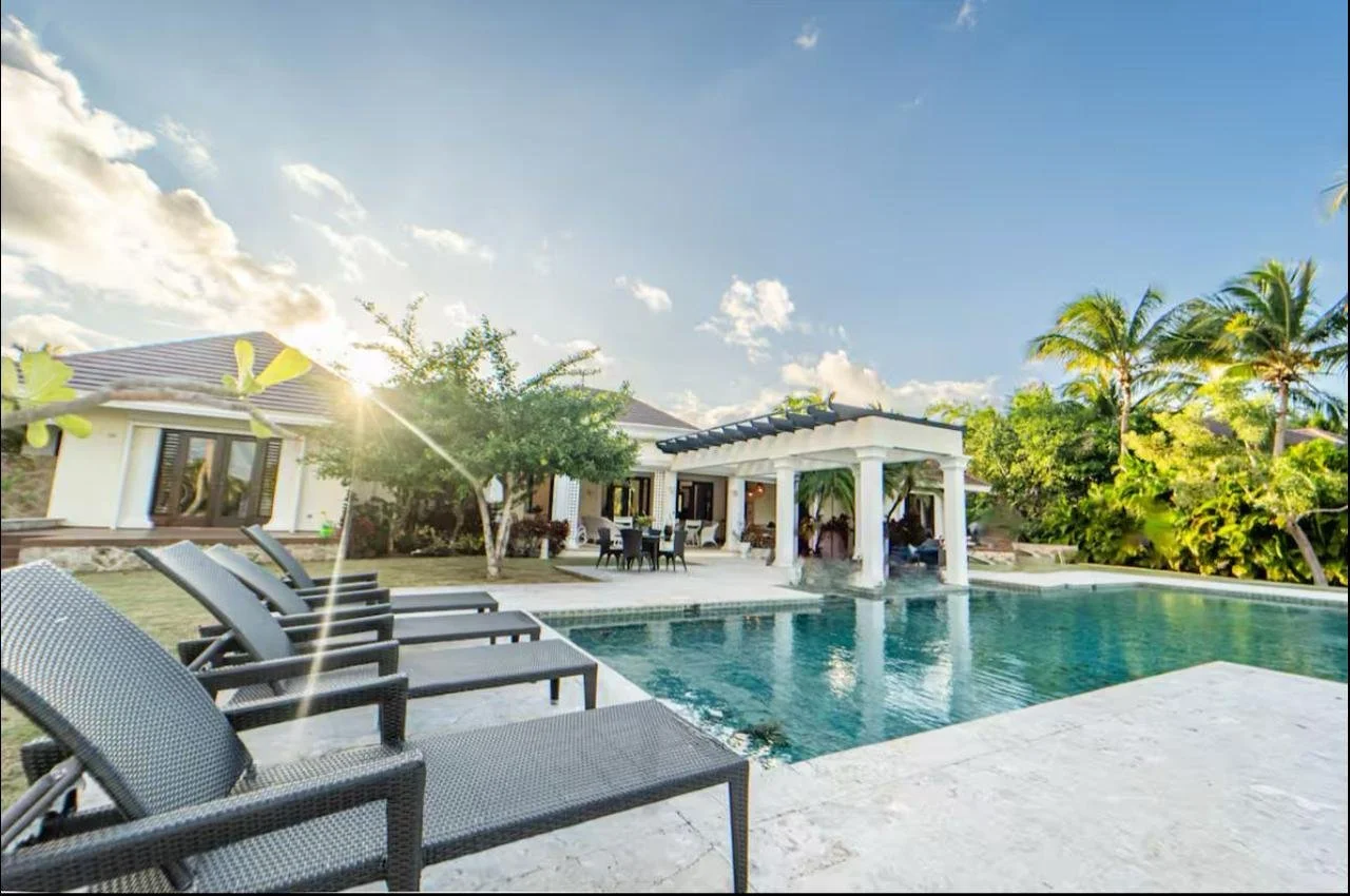 Piscina con tumbonas, palmas, árboles y una casa moderna con pérgola al atardecer