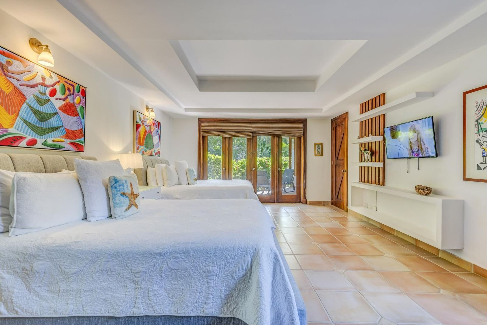 Habitación con cuatro camas de matrimonio con ropa de cama blanca, decorada con arte colorido en las paredes, con puertas de salida a una terraza con sillas, paredes blancas, suelo de baldosas y ventana de madera.