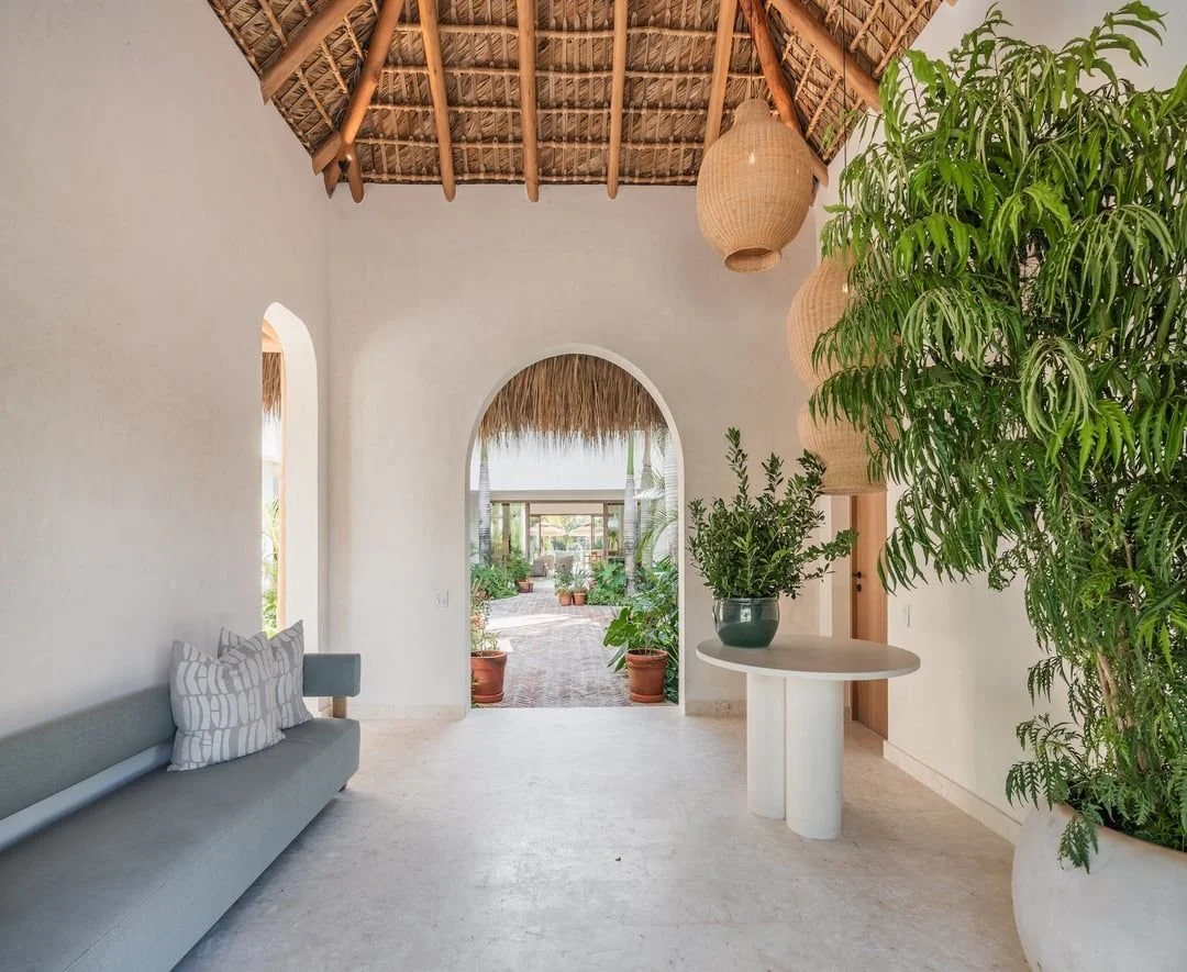 Interior de una casa con paredes blancas, piso de piedra, techo de palma y decoración rústica. Hay un sofá gris con cojines a la izquierda, plantas verdes en macetas a la derecha, y lámparas de mimbre colgantes. Se ve un arco que lleva a un patio exterior con más plantas.
