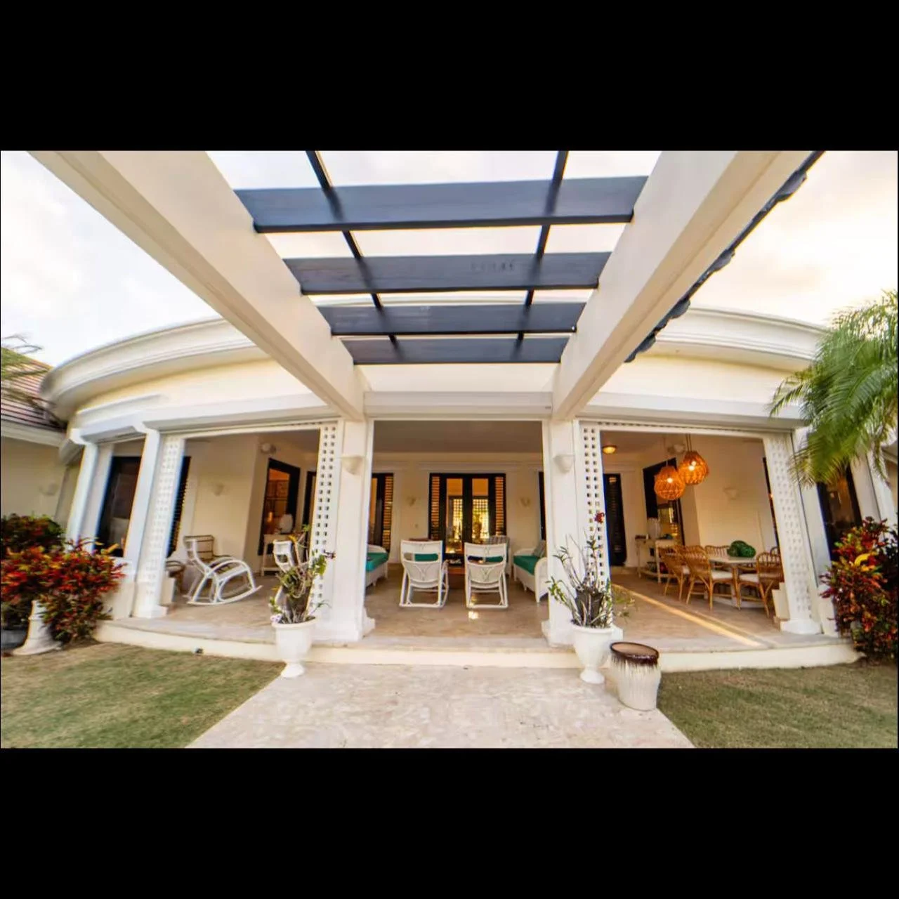 Fachada de casa moderna con área de terraza, columnas blancas, muebles de madera y plantas decorativas.