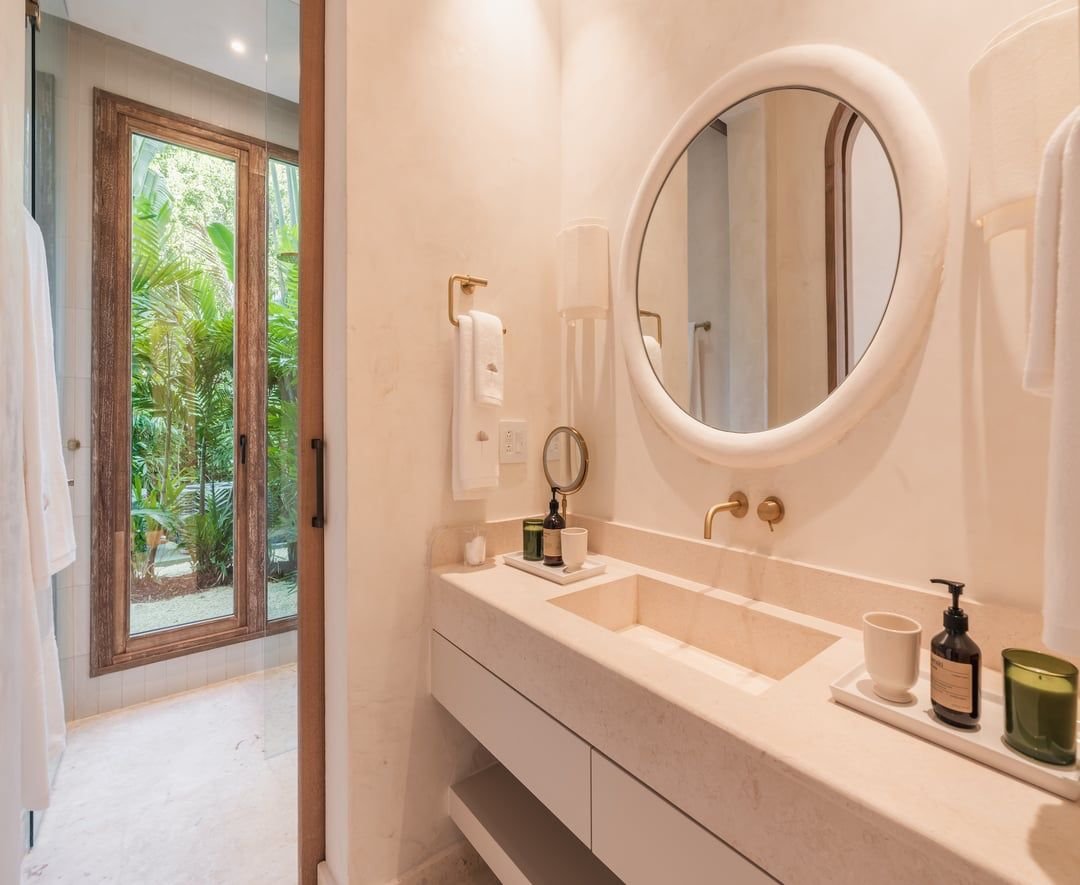 Lavabo con espejo redondo, ducha con puertas de vidrio y vista a un jardín con plantas verdes.