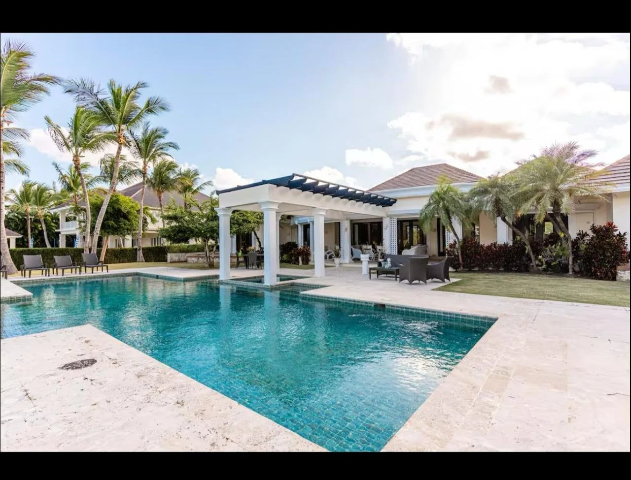 Piscina en un patio de casa con sillas y sombra, rodeada de palmeras y casas en un entorno tropical.