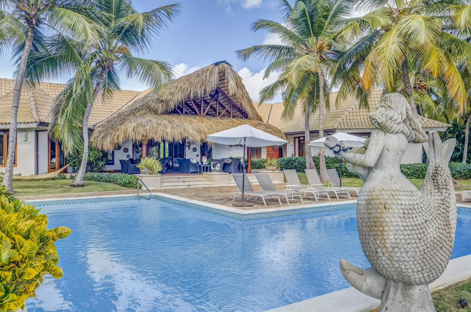 Piscina en un área de resort con palmeras altas, tumbonas y sombrillas, y una vivienda con techo de paja en el fondo.