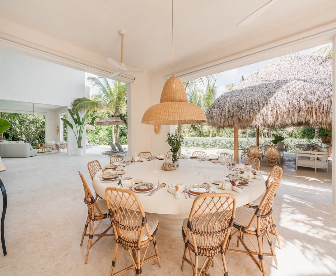 Mesa redonda decorada para una comida, en un espacio abierto y luminoso con plantas y muebles de exterior.
