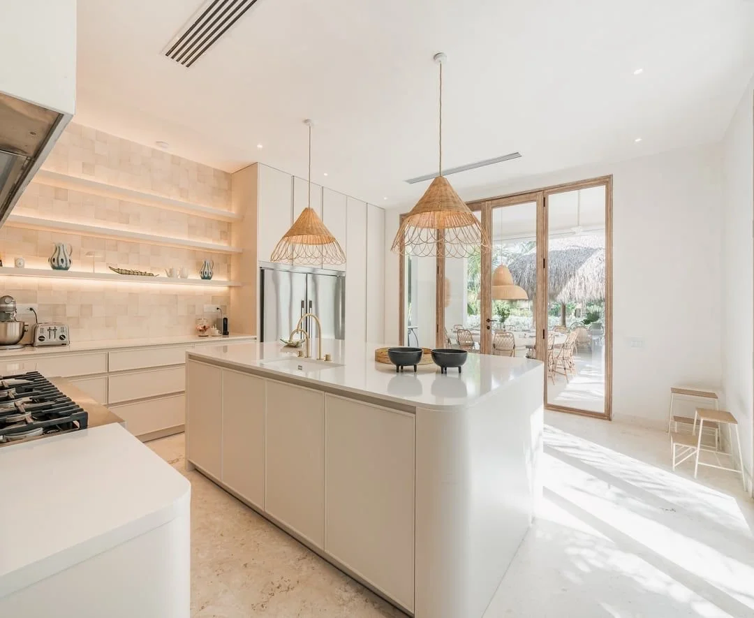 Cocina moderna y luminosa con isla central blanca, lámparas de mimbre, paredes de azulejo beige, refrigerador de acero inoxidable y salida a una terraza con sombra.