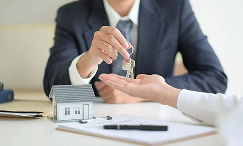 Persona entregando llave a otra en una oficina, con una maqueta de una casa y documentos sobre la mesa.