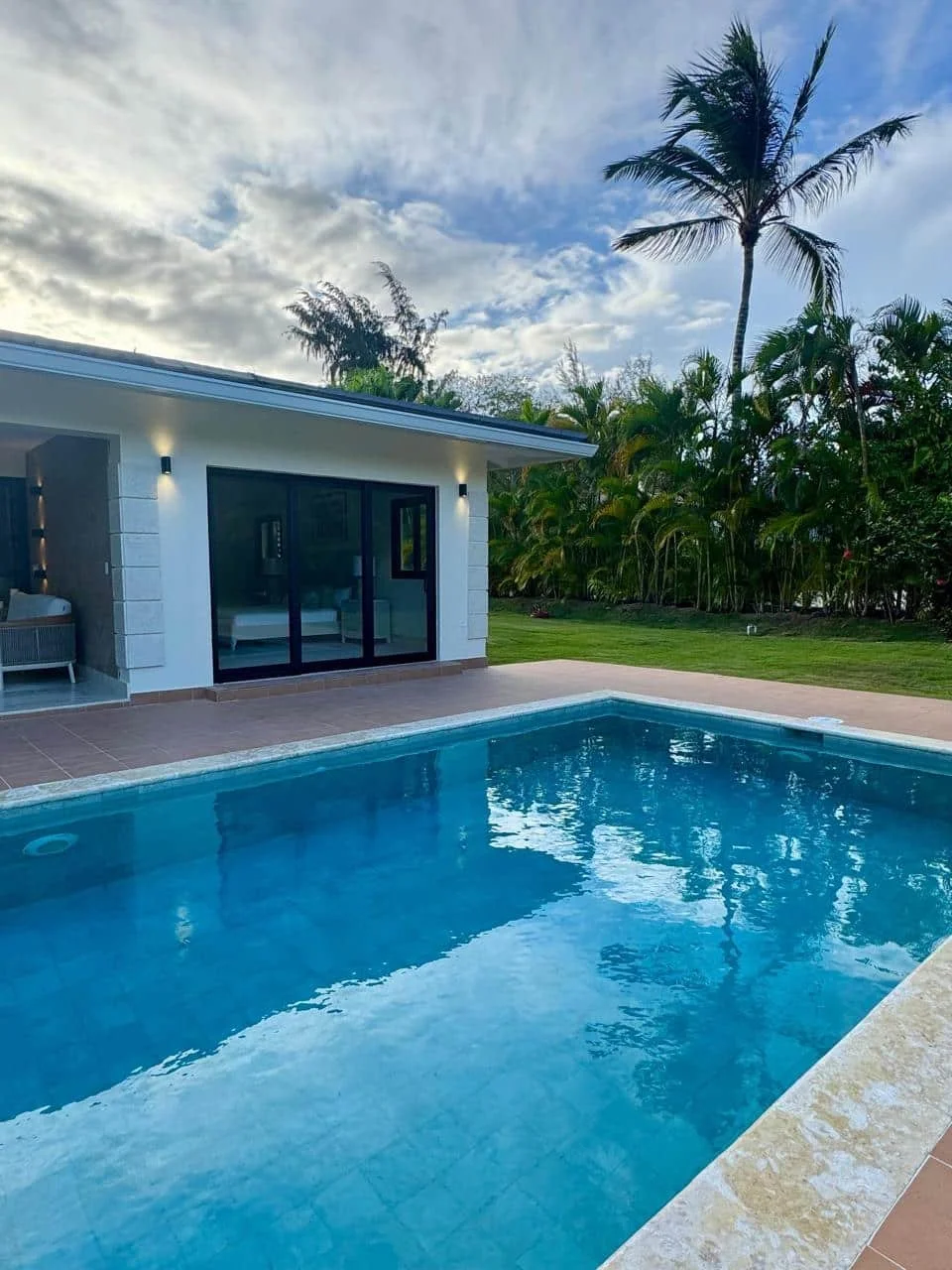 Piscina con borde de piedra, casa moderna con puerta de acceso y ventanas de vidrio, palmeras y plantas tropicales, cielo parcialmente nublado.