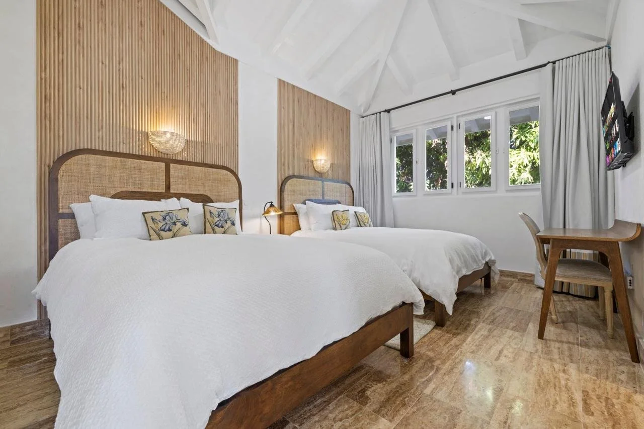 Habitación con dos camas grandes, cabeceras de mimbre, paredes decoradas con paneles de madera, iluminación de pared de estilo decorativo, ventana con cortinas blancas, piso de madera, televisor en la pared y mesa de madera con silla.