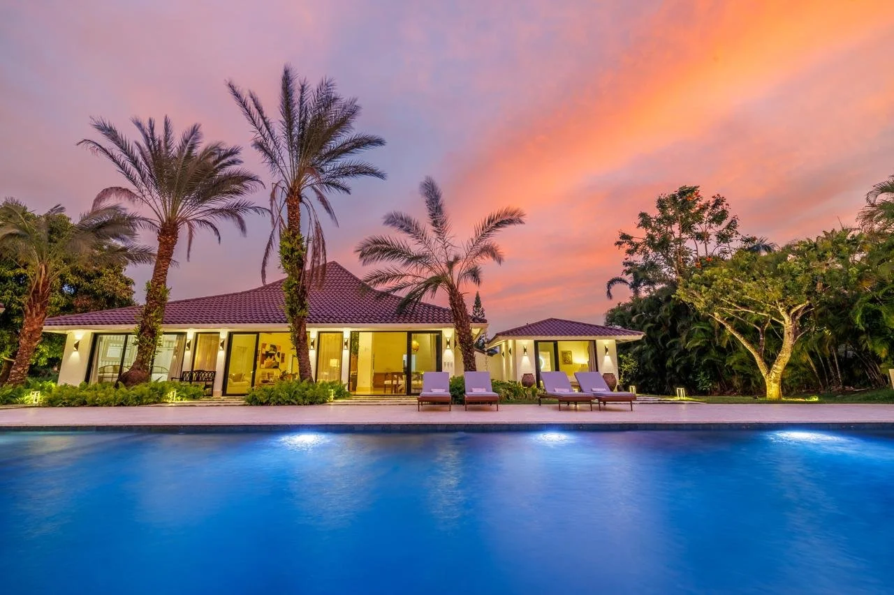 Casa moderna con piscina al atardecer, rodeada de palmeras y árboles.