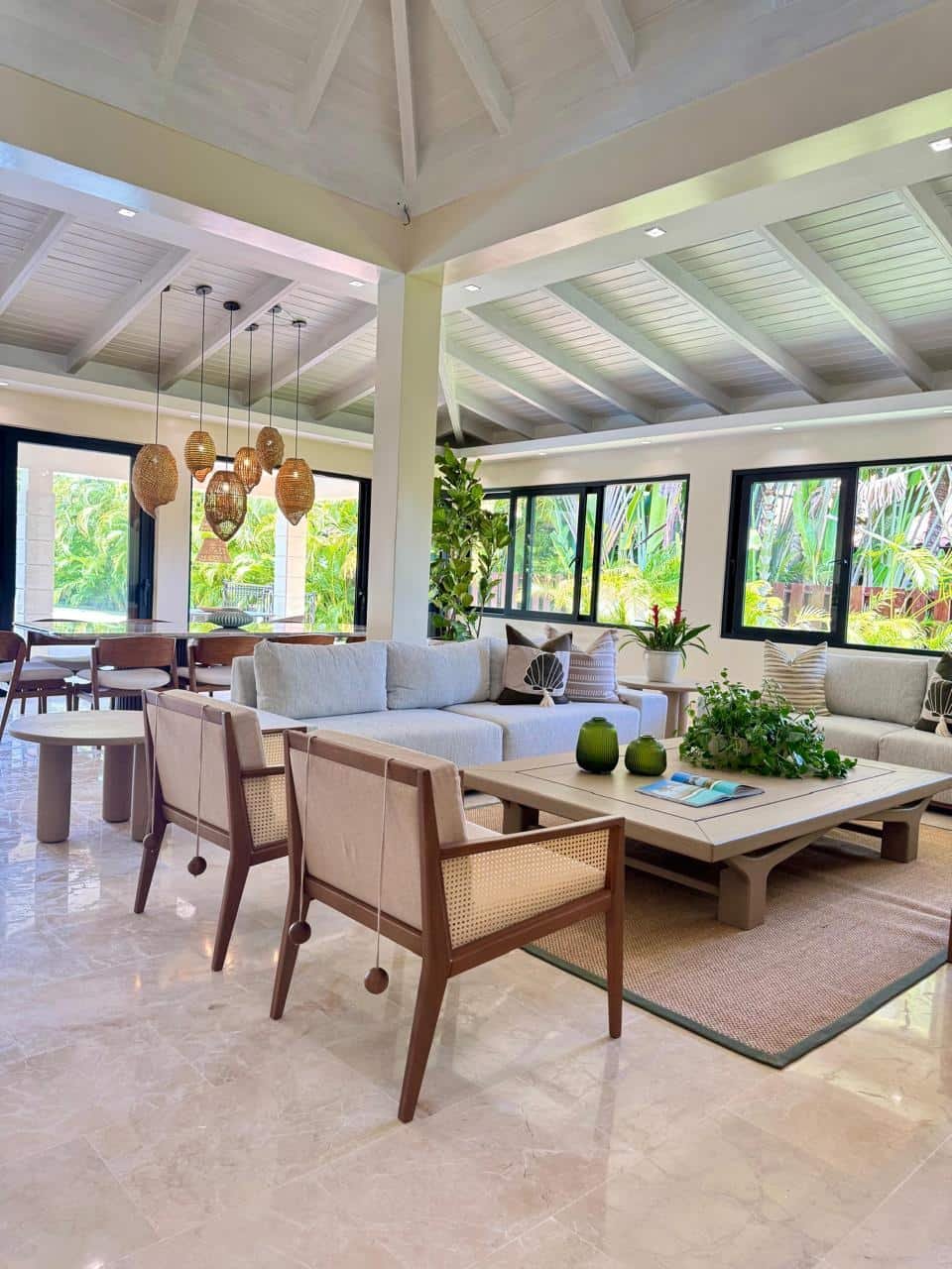 Sala de estar luminosa y moderna con sillones blancos, mesas de madera, plantas y ventanas grandes con vista a naturaleza tropical.