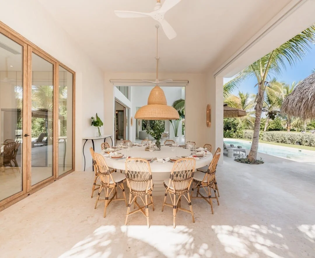 Comedor con mesa redonda y sillas de mimbre, decorado con plantas y vistas a una piscina y palmeras exteriores.