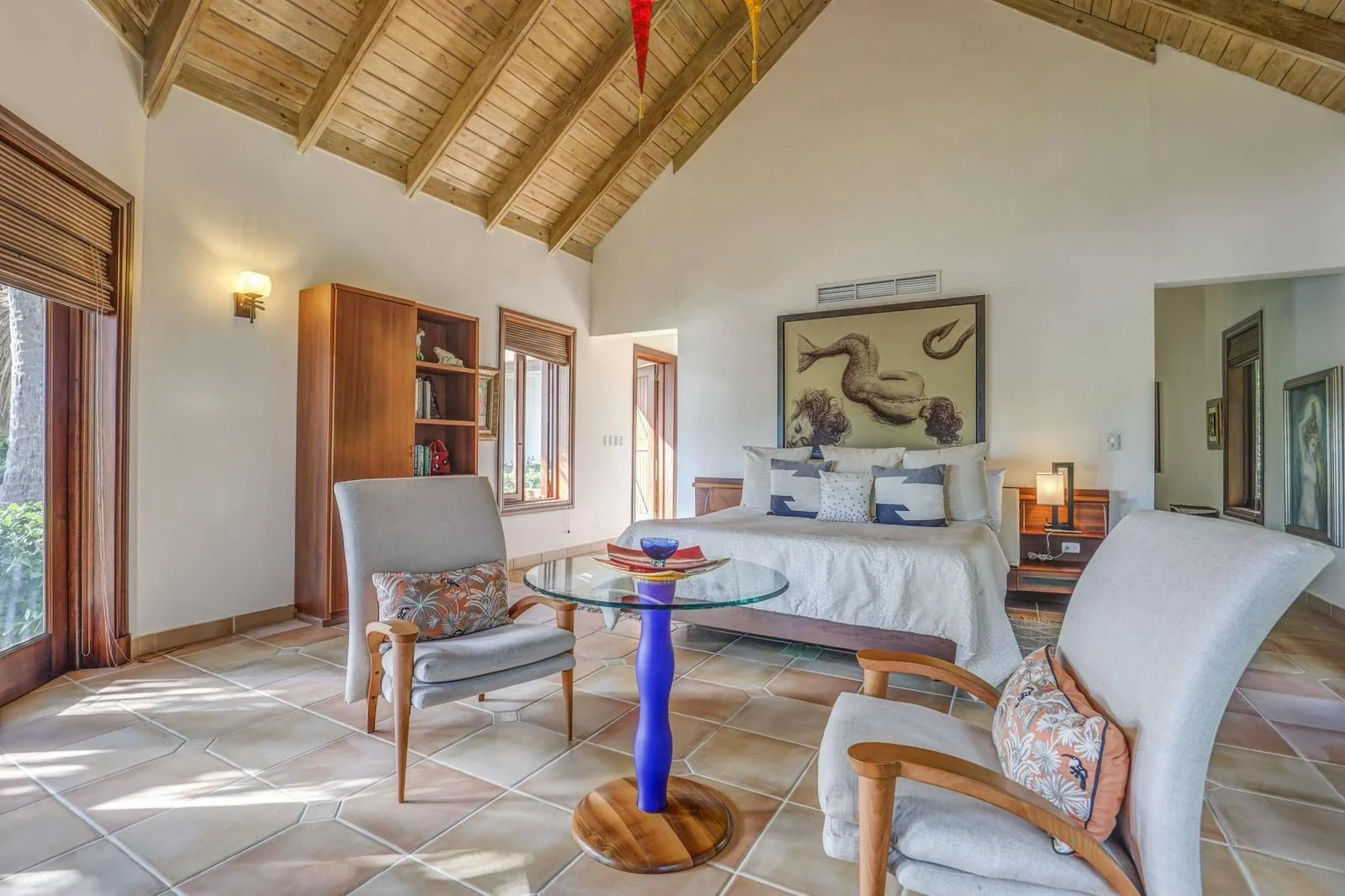 Sala de estar acogedora con cama grande, sillas de madera con cojines, mesa de centro de vidrio y techo de madera, ventanas grandes que dejan entrar luz natural, paredes blancas y decoración artística en la pared.