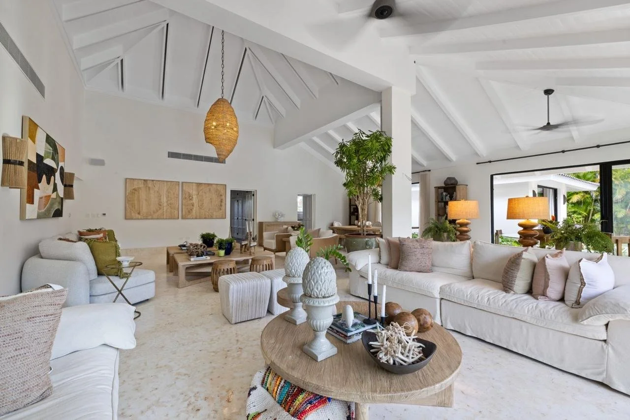 Sala de estar moderna con muebles blancos, lámparas de mesa con pantallas de color beige, planta grande en medio, paredes blancas, decoración en colores neutros y ventanas grandes con vista al jardín.