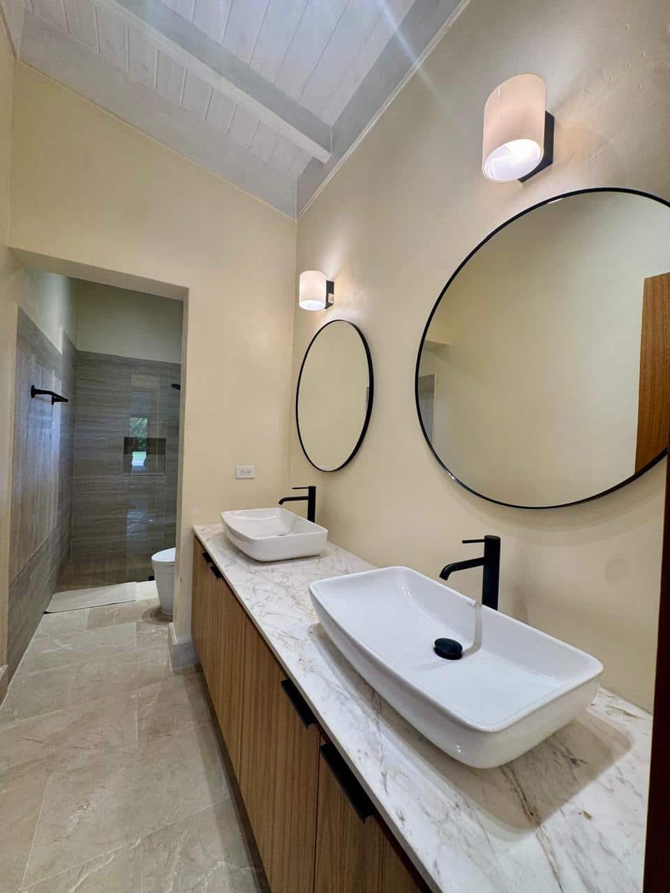 Lavabo doble con encimera de mármol, dos espejos redondos, paredes claras, iluminación moderna, piso de piedra beige, baño en el fondo con ducha y váter.