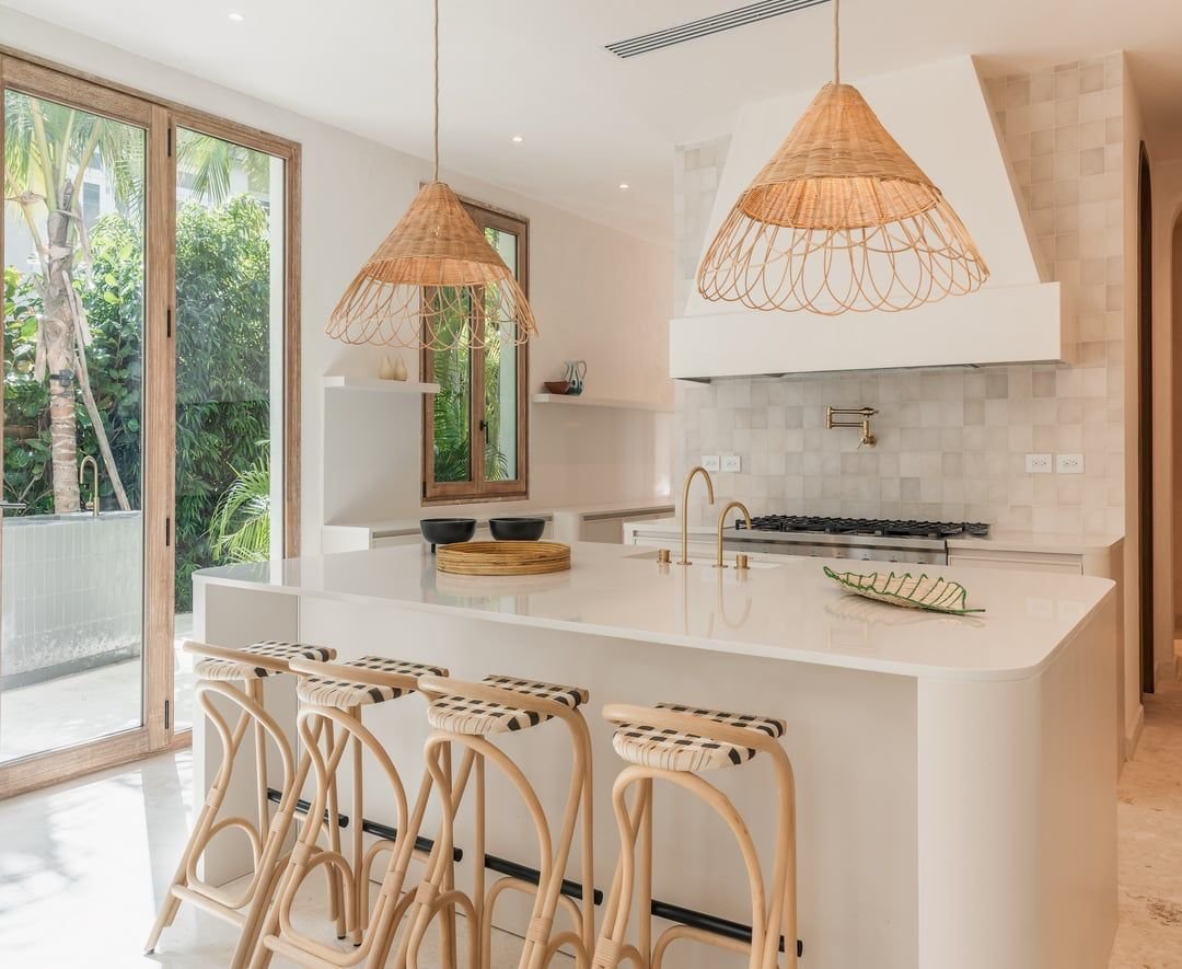 Cocina moderna con isla blanca y asientos de madera con cojines de rayas. Ventanas grandes con vistas a vegetación. Lámparas de techo de mimbre y detalles en tonos neutros.