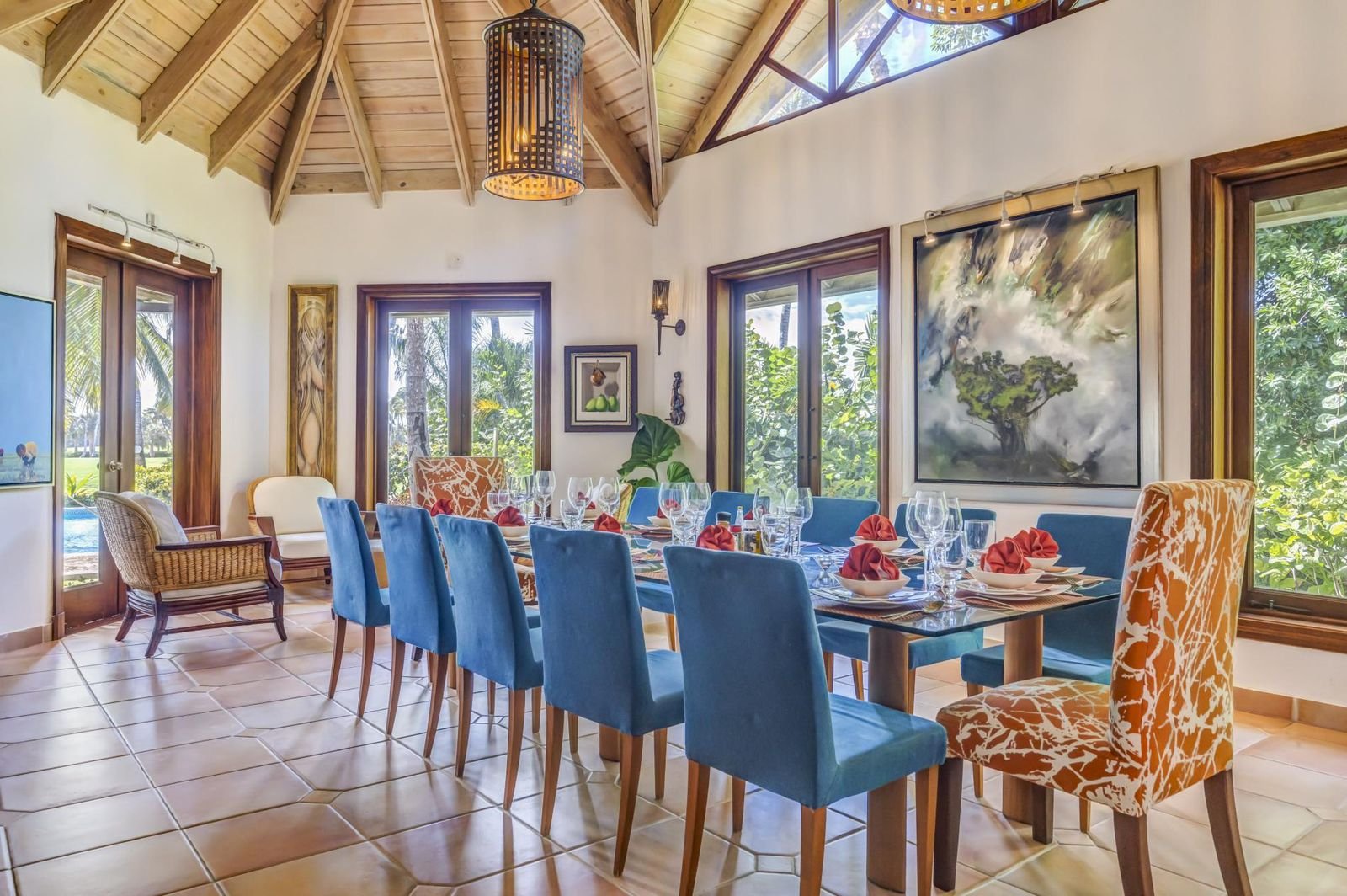 Comedor decorado con sillas azules y naranjas, mesa con copas, platos y manteles, en una habitación con paredes blancas, ventanas grandes con vista a un jardín, pintura en la pared y techo de madera
