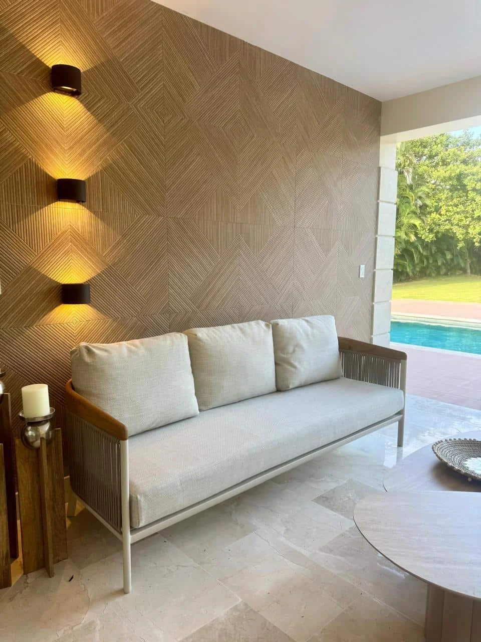 Sofá blanco con cojines en un espacio con pared decorativa y vista a una piscina exterior y jardinería