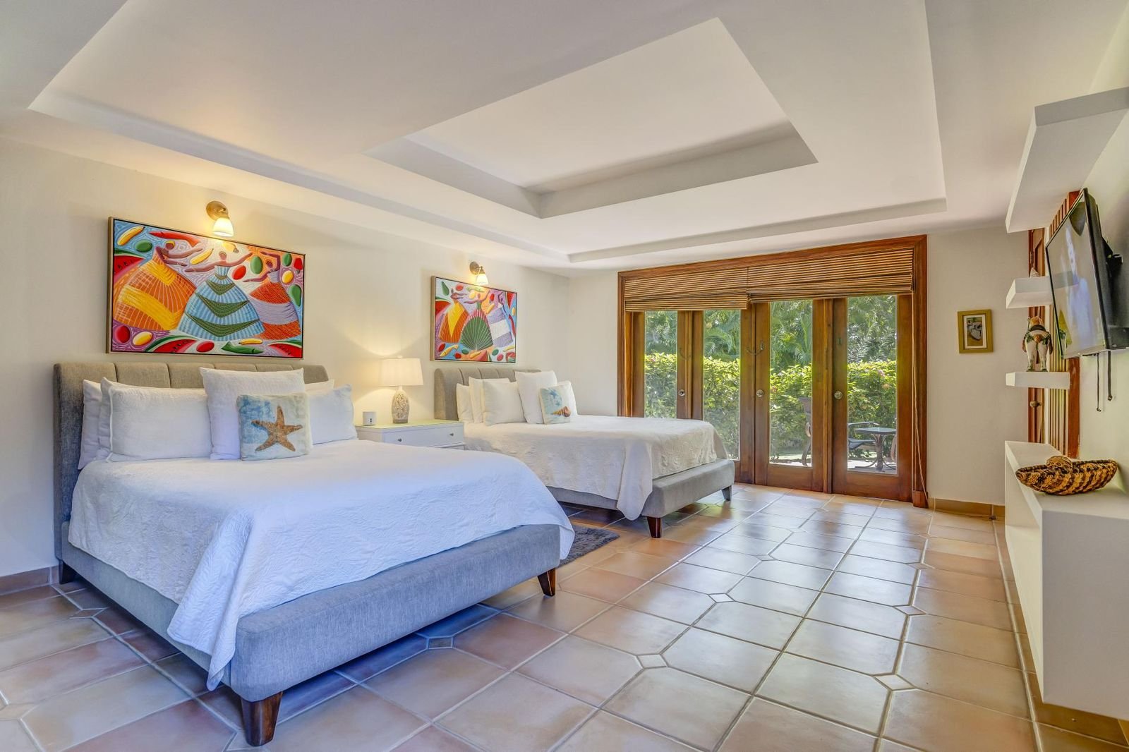 Habitación de hotel con dos camas grandes con ropa blanca, arte colorido en las paredes, un ventanal con vista a la vegetación, muebles blancos y decoración tropical.