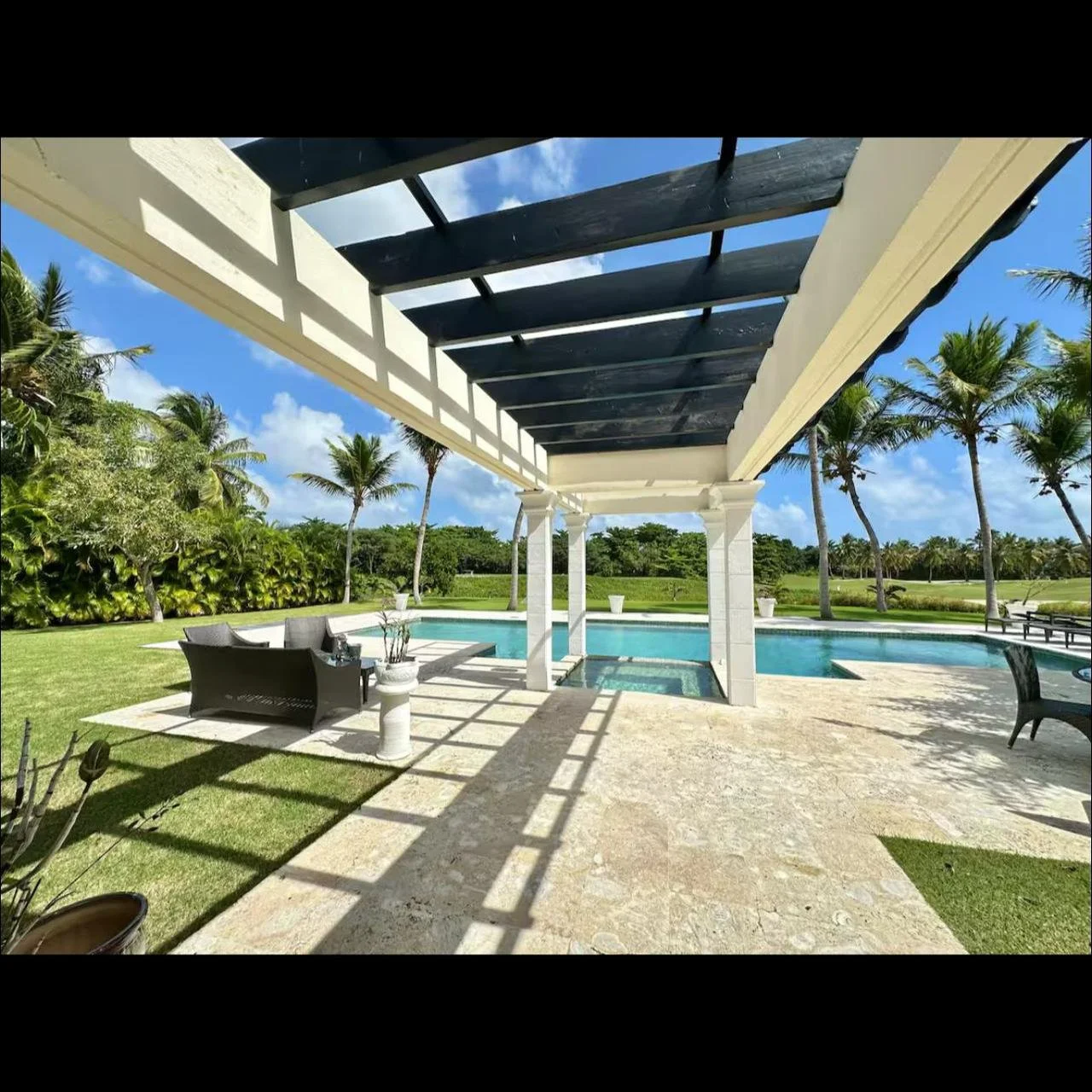 Vista desde una estructura de patio con sombra sobre una piscina y césped, rodeada de palmeras y cielo despejado.