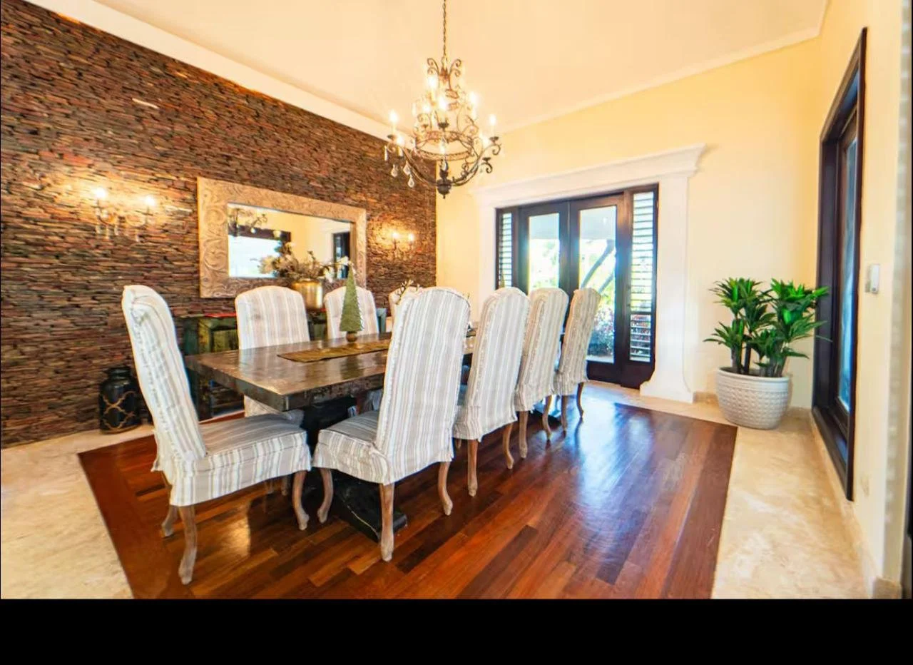Comedor con paredes de ladrillos, mesa de madera y sillas con tapizado a rayas, lámpara de araña y plantas decorativas en un ambiente bien iluminado.