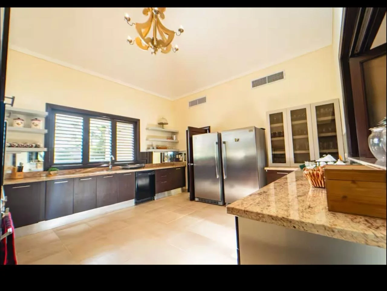 Cocina moderna con gabinetes oscuros, encimeras de granito, refrigeradores de acero inoxidable, estantes blancos, ventana con persianas, lámpara de araña y suelo de baldosas beige.