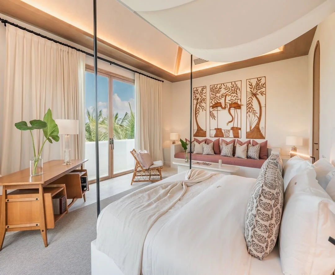 Habitación moderna con vista al exterior, decoración en tonos claros, un ventanal con cortinas blancas, arte en la pared con motivos de naturaleza, y muebles de madera con plantas.