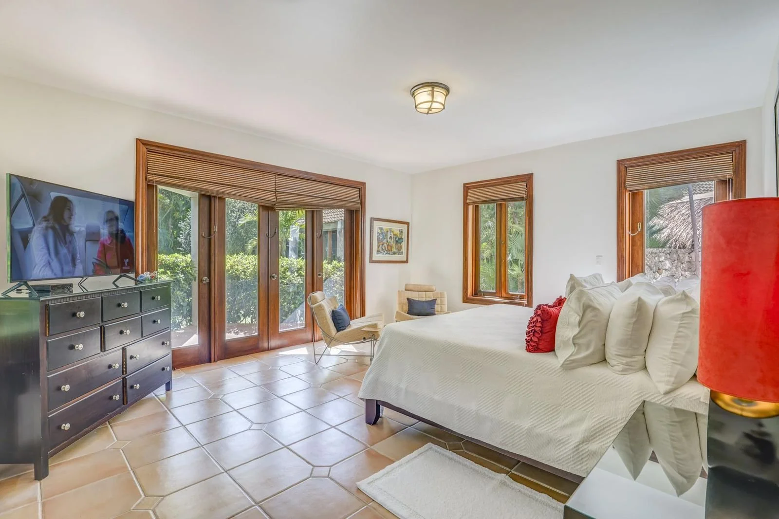 Habitación con una cama blanca con cojines rojos y blancos, televisión en un mueble negro, ventanas con vistas a un jardín verde, sillones y cuadros decorativos.